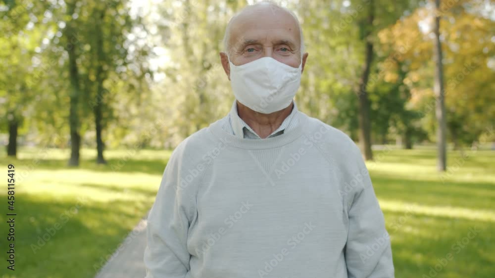 Slow motion portrait of senior man wearing medical face mask standing in summer park alone and looking at camera. Pandemic and people concept. Slow motion portrait of senior man wearing medical face mask standing in summer park alone and looking at camera. Pandemic and people concept.