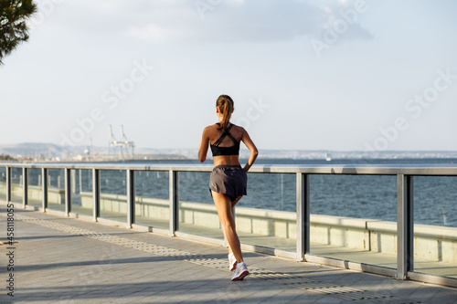 Fototapeta Naklejka Na Ścianę i Meble -  beautiful fitness girl in shorts and sports top running on the waterfront by the sea in sunny weather