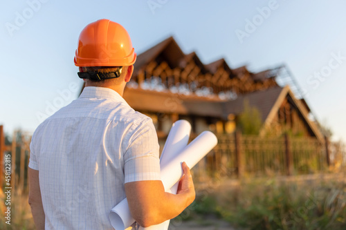 Young engineer in a yellow helmet with blueprints at a construction site in front of a building requiring reconstruction or repair
