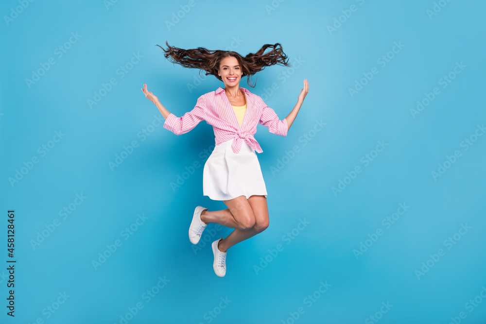 Photo of shiny charming young woman dressed white skirt smiling rising hands arms jumping high isolated blue color background Photo of shiny charming young woman dressed white skirt smiling rising hands arms jumping high isolated blue color background
