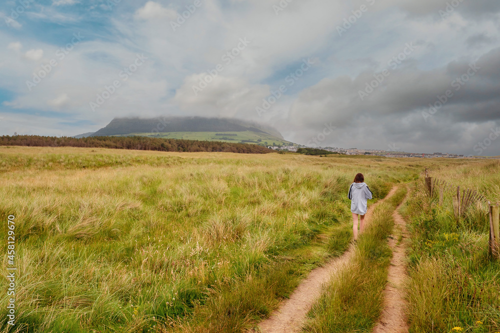 Young slim teenager girl walking on a small road towards Strandhill town, Knocknarea hill in cloudy sky in the background. County Sligo, Ireland. Irish landscape. Popular landmark.