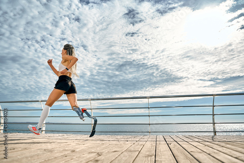 Take care of your body. Side view of a strong disabled woman in sportswear with a prosthetic leg is running on the bridge.