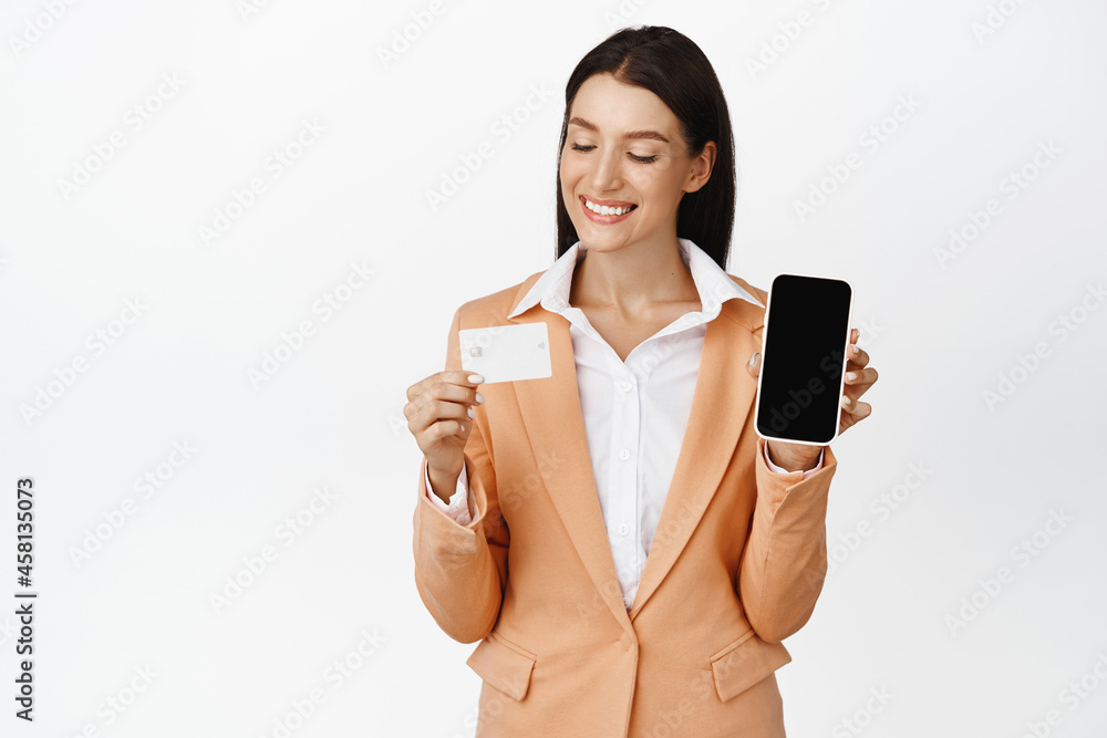 Smiling saleswoman in suit showing her phone screen, looking pleased at credit card, standing against white background