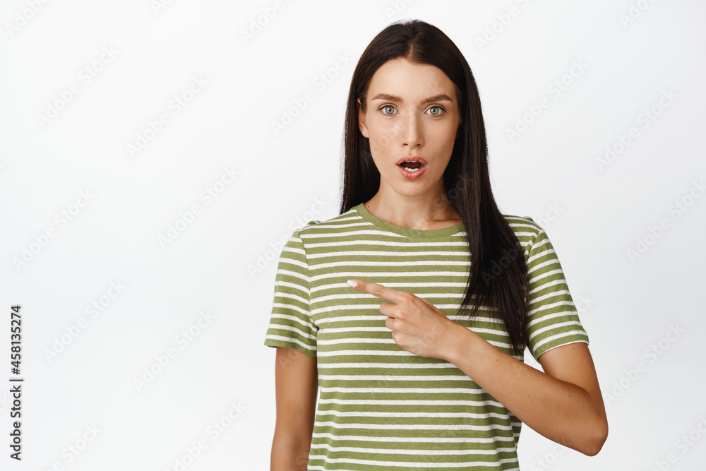 Amazed brunette woman pointing finger left, looking impressed at camera, wearing casual striped t-shirt Amazed brunette woman pointing finger left, looking impressed at camera, wearing casual striped t-shirt