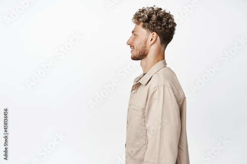 Profile shot of handsome blond guy standing in jacket and smiling, looking left side, standing against white background
