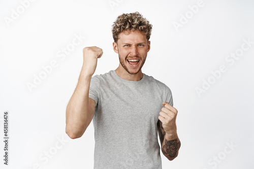Cheerful handsome young man in grey t-shirt, celebrating, scream from joy and making fist pumps, acheive goal and triumphing, white background photo