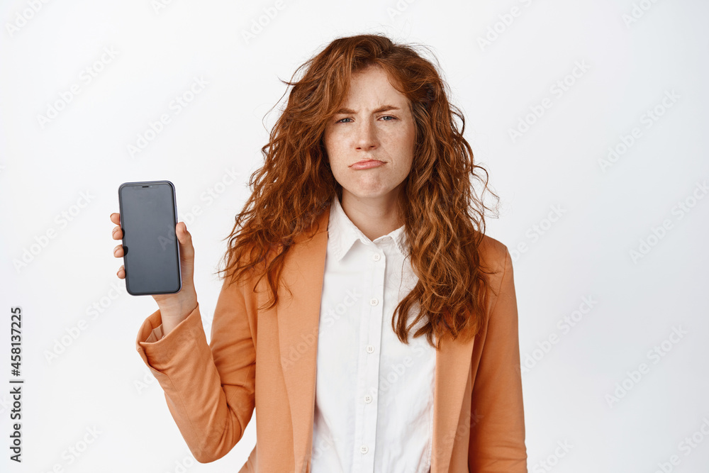 Naklejka premium Sad frowning business woman showing mobile phone screen and sulking, dislike smartphone app, standing in suit against white background