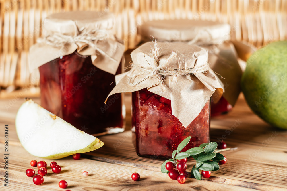 Jars of homemade lingonberry and pear jam with craft paper on lids on wooden table next to fresh lingonberries and pears Jars of homemade lingonberry and pear jam with craft paper on lids on wooden table next to fresh lingonberries and pears