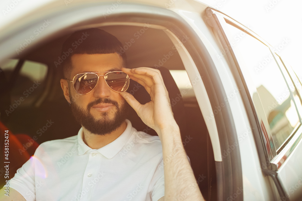 young man with beard wearing sunglasses smiles while driving a car