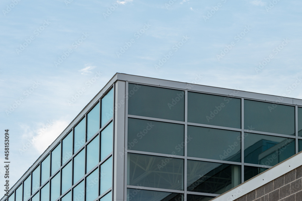 Exterior corner of a commercial building with glass windows reflecting the sky and a dark brown brick wall base with grey mortar. The background is a blue sky with white clouds on a sunny day. Exterior corner of a commercial building with glass windows reflecting the sky and a dark brown brick wall base with grey mortar. The background is a blue sky with white clouds on a sunny day.