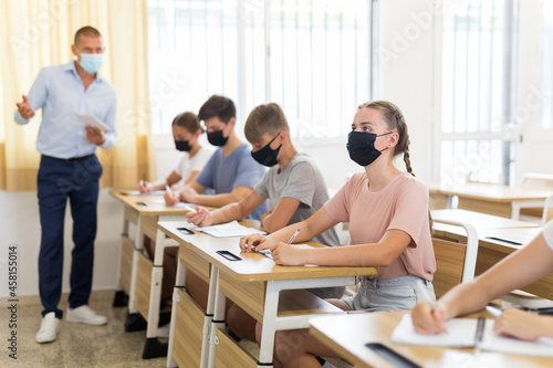 Teenage students in protective mask studying in classroom with teacher, writing lectures in workbooks