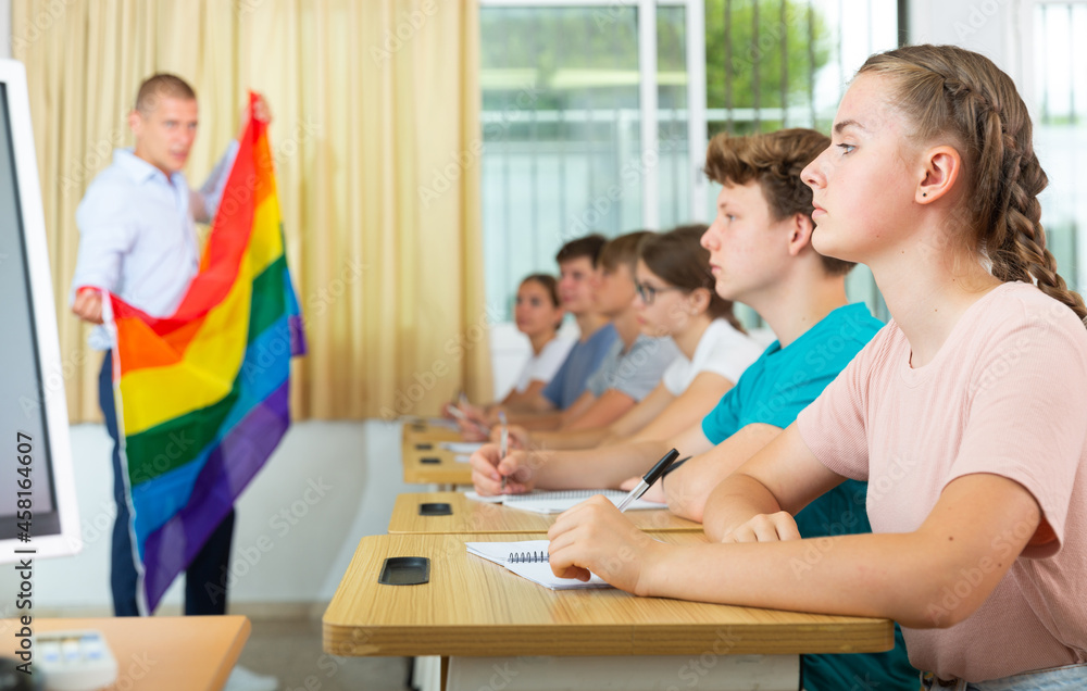 Fototapeta premium Focused teenage schoolgirl sitting at lesson with classmates, listening young teacher talking about LGBT community and showing rainbow flag