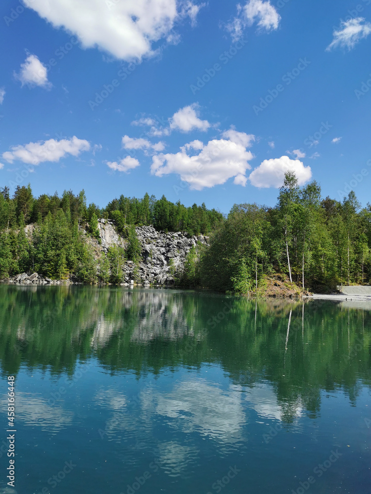 The shore of the lake Light with turquoise water, which reflects the trees and the sky with clouds, in the mountain park Ruskeala.