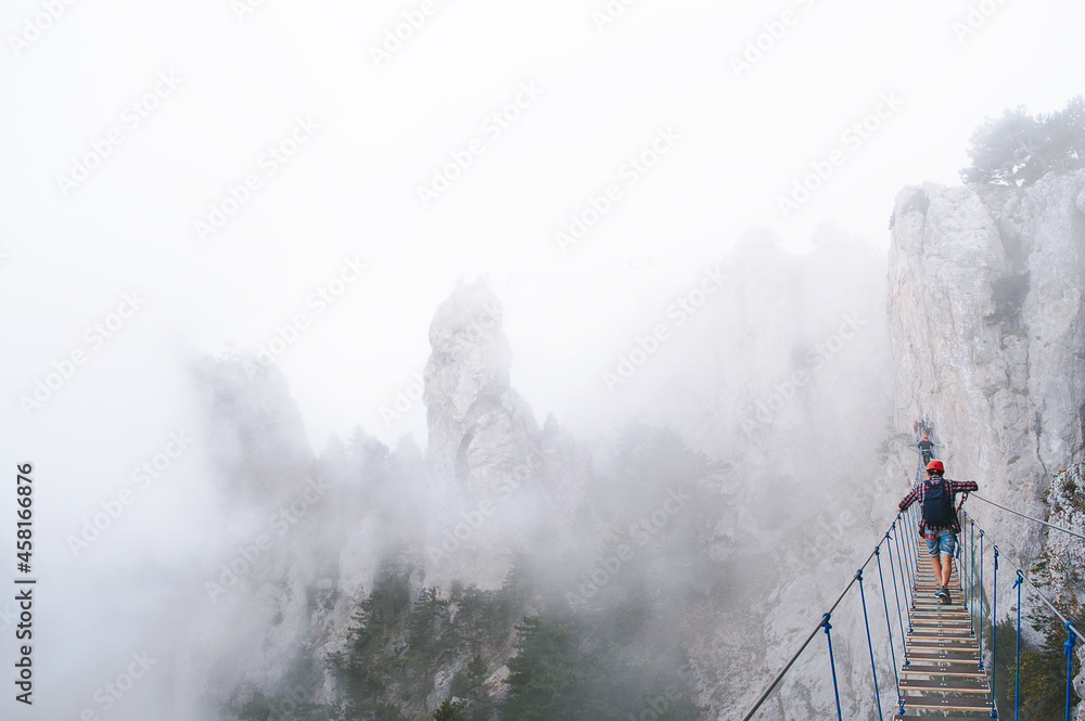 A young man in mountaineering equipment walks uphill on a suspension bridge in the fog. VIA Feratta - Heavenly trail. Suspension bridge on Mount Ai-Petri, Yalta, Crimea. Sea coast in Crimea.  - obrazy, fototapety, plakaty 