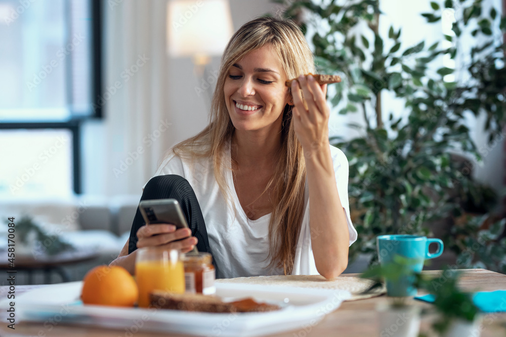 Smiling young woman enjoying breakfast while using her mobile phone in the living room at home.