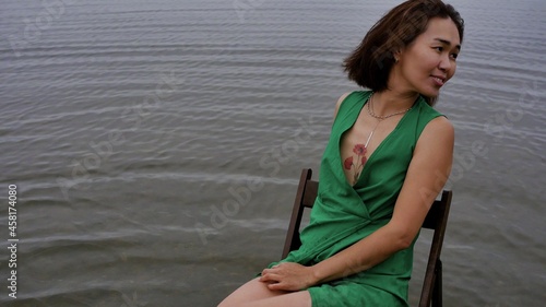 A beautiful Asian woman sits on a chair in the water by the ocean in the early morning and splashes salt water with camla, laughs and poses for a photographer. Happy woman in green dress sits on a photo