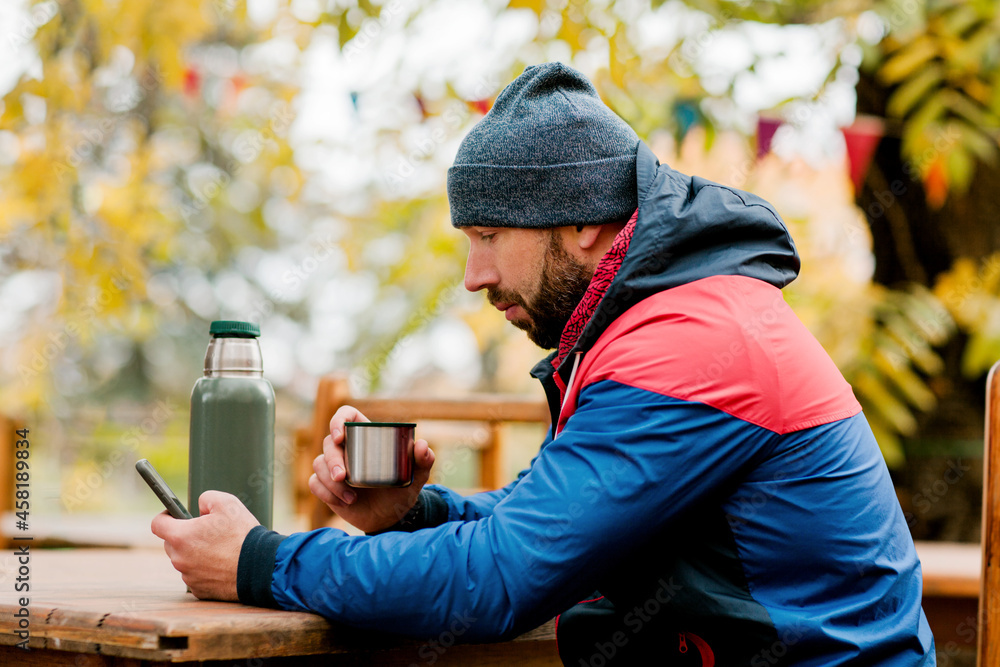 A young man using a mobile phone is sitting outdoors in a cafe or restaurant on an autumn day, drinking a hot drink, resting while walking A young man using a mobile phone is sitting outdoors in a cafe or restaurant on an autumn day, drinking a hot drink, resting while walking