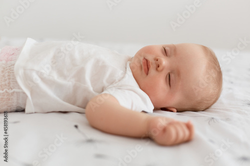 Side view of charming sleeping female kid wearing white t shirt lying on bed on white sheet with closed eyes, posing indoor at home, happy carefree childhood.