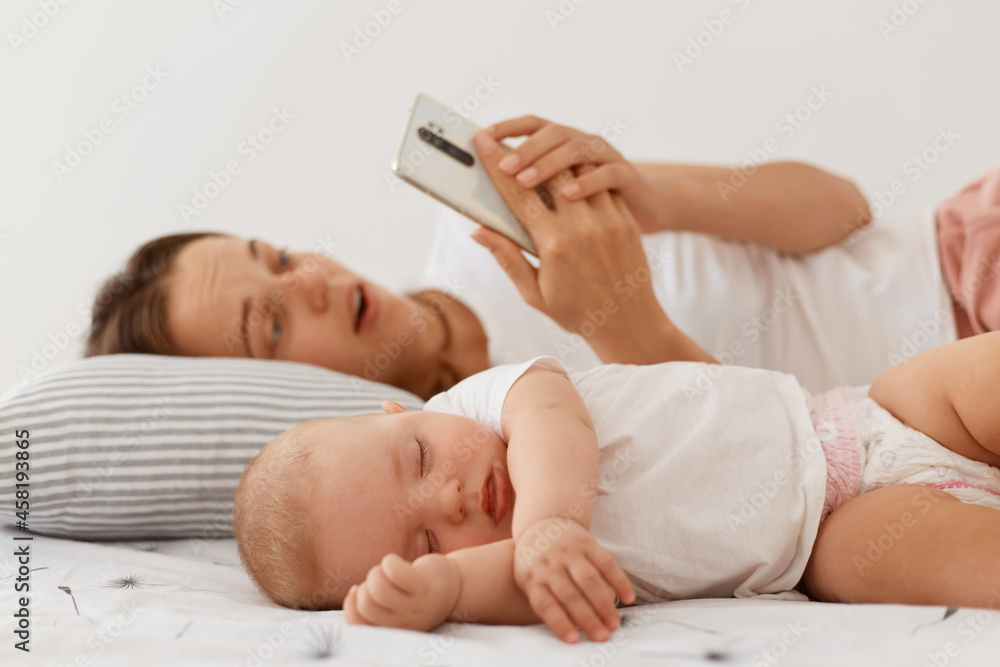 Portrait of surprised female wearing white t shirt and holding smart phone, looking at smartphone screen with open mouth and astonishment, lying with sleeping baby daughter.