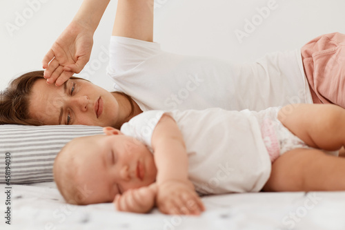 Portrait of dark haired female wearing white casual t shirt lying on bed with little baby daughter, posing indoor, woman looking at her infant girl with tired expression.