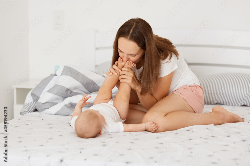 Indoor shot of happy positive woman wearing white t shirt and shorts kissing daughter's feet, playing with baby at home, sitting on bed at home, happy motherhood. Indoor shot of happy positive woman wearing white t shirt and shorts kissing daughter's feet, playing with baby at home, sitting on bed at home, happy motherhood.