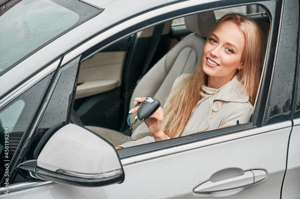 Portrait of beautiful blond girl sitting in automobile with lowered window and holding car keys. Successful smiling business lady with happy face expression showing keys while driving her white auto.