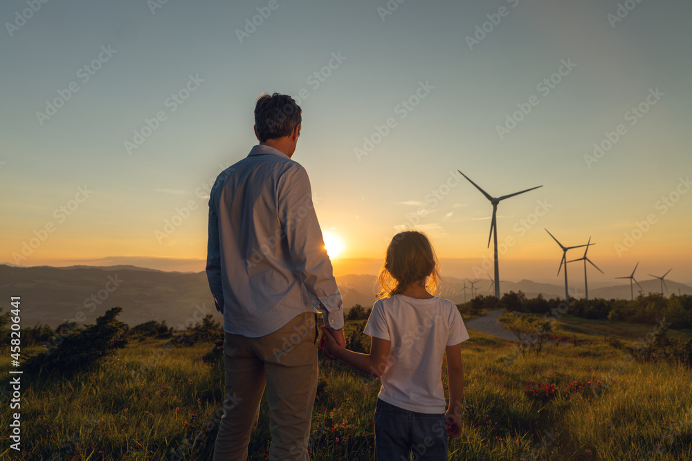 Cinematic shot of carefree young father engineer keeping his daughter for hand and looking on windmill field at sunset. Concept of renewable energy, love for nature, family, electricity, green, future - obrazy, fototapety, plakaty 