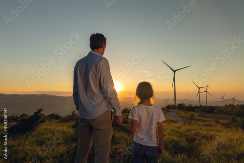 Cinematic shot of carefree young father engineer keeping his daughter for hand and looking on windmill field at sunset. Concept of renewable energy, love for nature, family, electricity, green, future photo