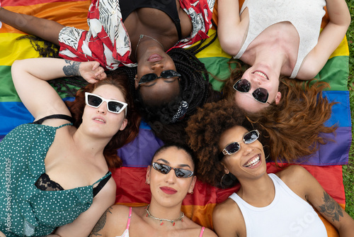 Cinematic shot of girl friends of different ethnicities with sunglasses while lying on LGBT rainbow flag in park. Concept of friendship, homophobia, diversity, equality, freedom, liberty, multiethnic. photo