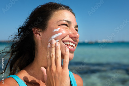 Canvas Print Close up of happy smiling young woman is applying sunscreen or sun tanning lotion on face to take care of her skin on seaside beach during holidays vacation trip