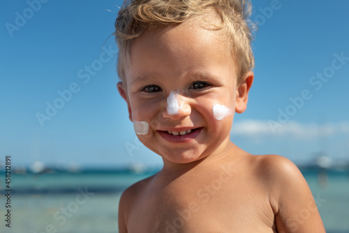 Foto Cute happy little toddler boy is smiling in camera with protective sunscreen or sunblock lotion on his face applied by his mother to take care of skin on seaside beach during family holidays vacation