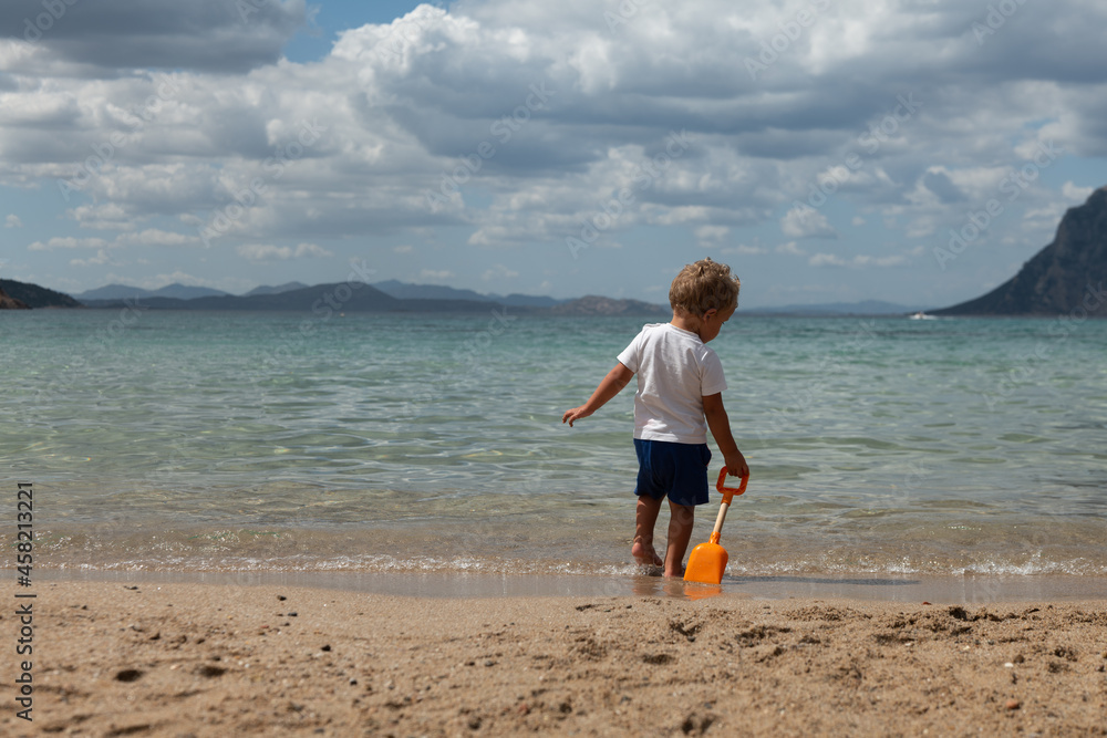Authentic back shot of little toddler baby boy playing with scapula toy for sand on scenic seascape during family holidays vacation trip. Concept of travel, tourism, childhood, happiness and freedom.