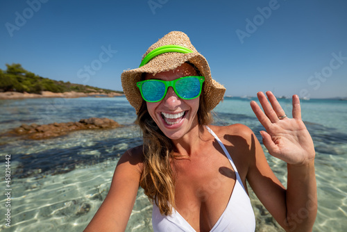 A happy young smiling female tourist in white bikini, straw hat and sunglasses making selfie or technology video call with to friends or family in sea during holidays vacation trip. Concept of roaming