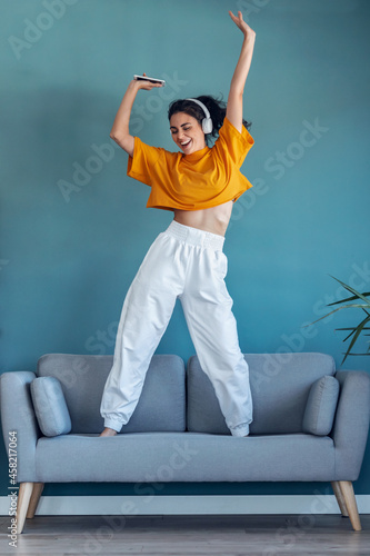 Funny woman listening to music with smartphone while dancing on the couch in the living room at home.