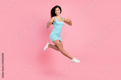 Full size profile photo of funny young lady jump show thumb up wear blue dress sneakers isolated on pink color background