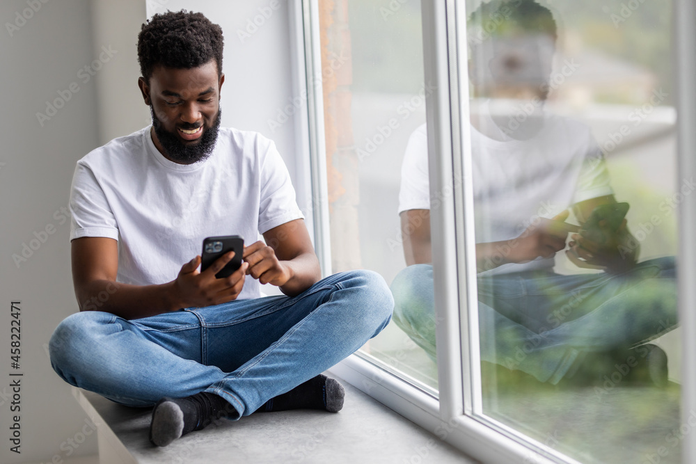 Handsome african man sits home on windowsill, reads text messages, sends email, uses his phone and fast internet connection. Handsome african man sits home on windowsill, reads text messages, sends email, uses his phone and fast internet connection.