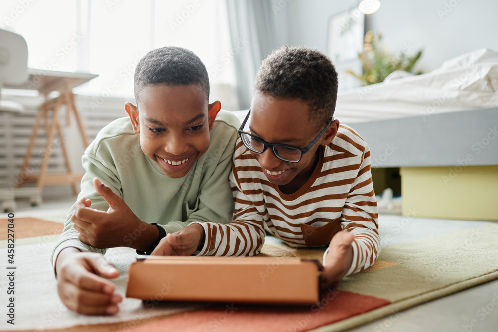 Portrait of two teenage African-American boys using digital tablet together while lying on floor in cozy room, copy space Portrait of two teenage African-American boys using digital tablet together while lying on floor in cozy room, copy space