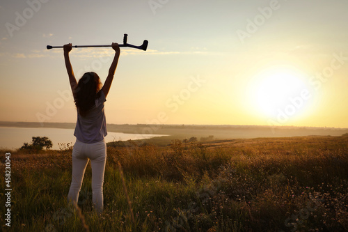 Woman raising hands with elbow crutch up to sky outdoors at sunrise, back view. Healing miracle