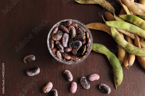Top down view of scarlet runner bean in a small crystal glass bowl on a dark surface. In the background there are green and yellow hulls and beans.