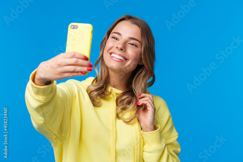 Close-up portrait tender cute blond girl in yellow hoodie, holding mobile phone, taking selfie with smartphone posing adding photo filter in appllication, standing blue background photo