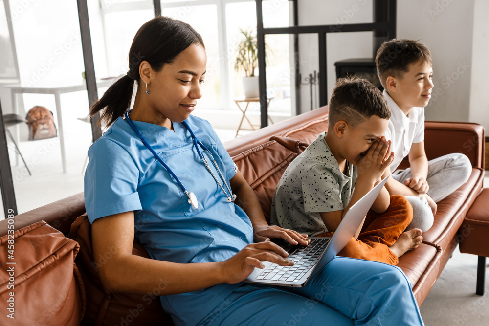 Fototapeta premium Black woman doctor using laptop while sitting with her sons on couch