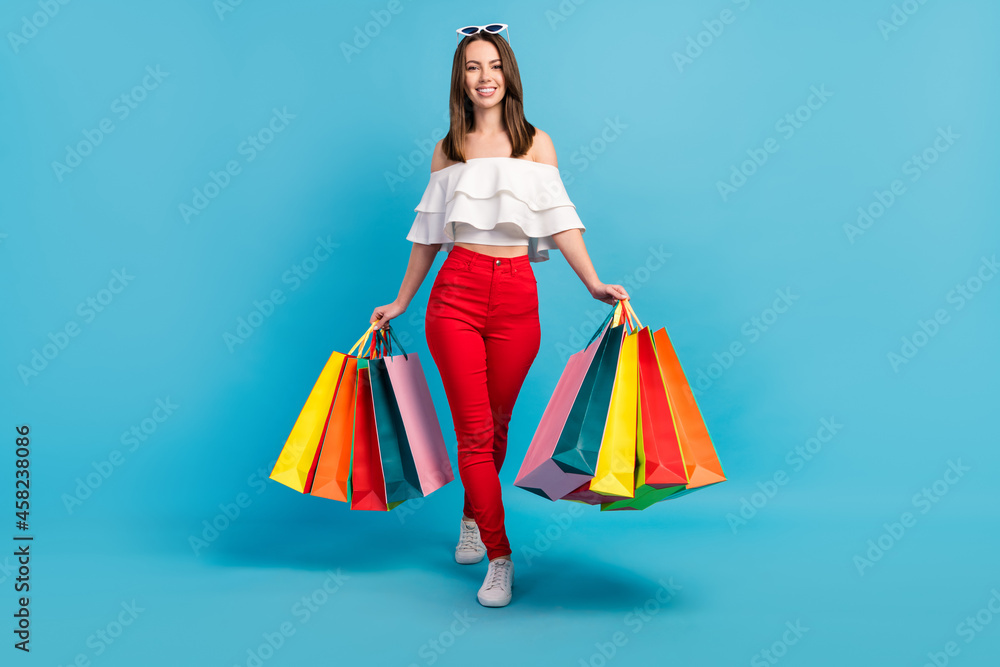 Photo of sweet shiny lady wear off-shoulders shirt dark eyewear walking holding shopping bags smiling isolated blue color background Photo of sweet shiny lady wear off-shoulders shirt dark eyewear walking holding shopping bags smiling isolated blue color background