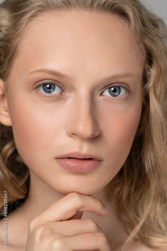 Obraz premium Close up of beauty woman face portrait with blue eyes and curly hair. Spa model girl with perfect fresh clean skin. Blond female looking at camera and touching her chin. Youth and skin care concept. 