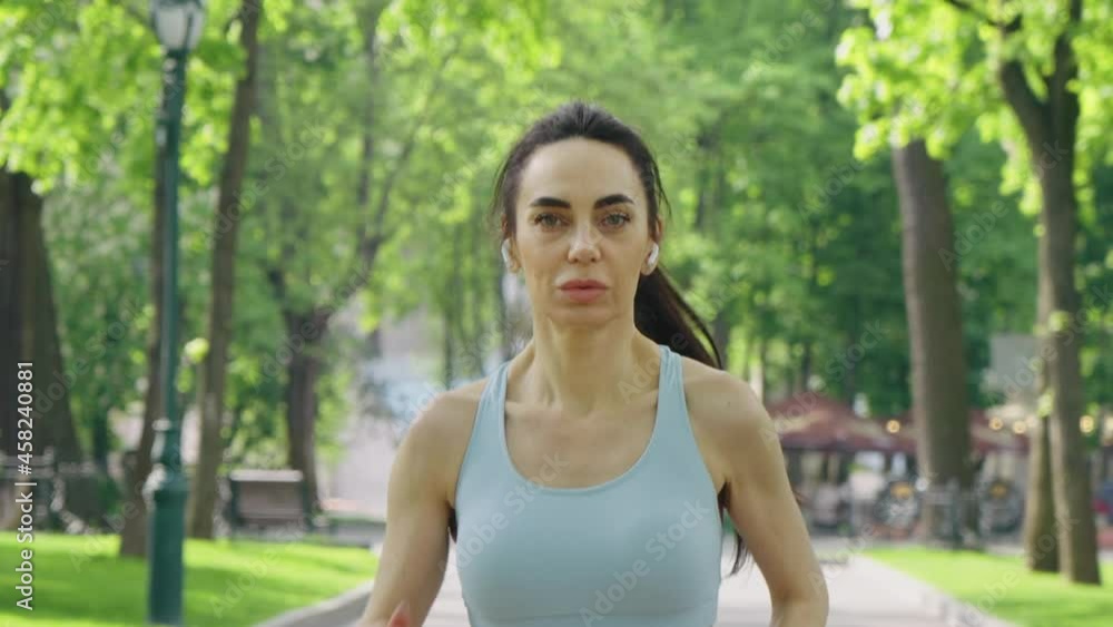 Woman with ponytail wearing blue top and wireless earphones jogging in park in slow motion. Tracking shot fit female looking at camera while training. Concept of healthy lifestyle Woman with ponytail wearing blue top and wireless earphones jogging in park in slow motion. Tracking shot fit female looking at camera while training. Concept of healthy lifestyle