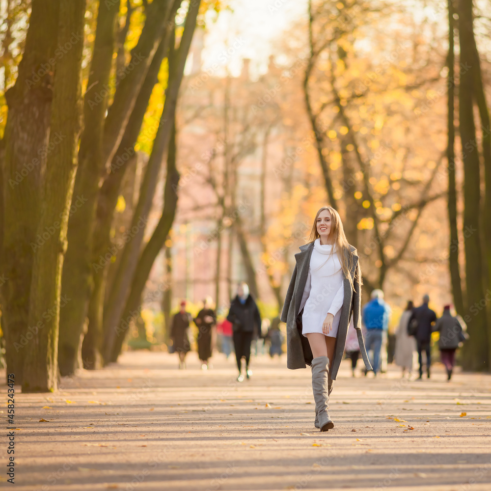 Fototapeta premium Happy young woman walks in the autumn park. An attractive woman with light brown hair and a gray coat is walking down the alley. Beautiful autumn mood. Fall season.