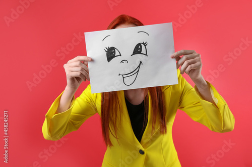 Woman hiding behind sheet of paper with happy face on red background