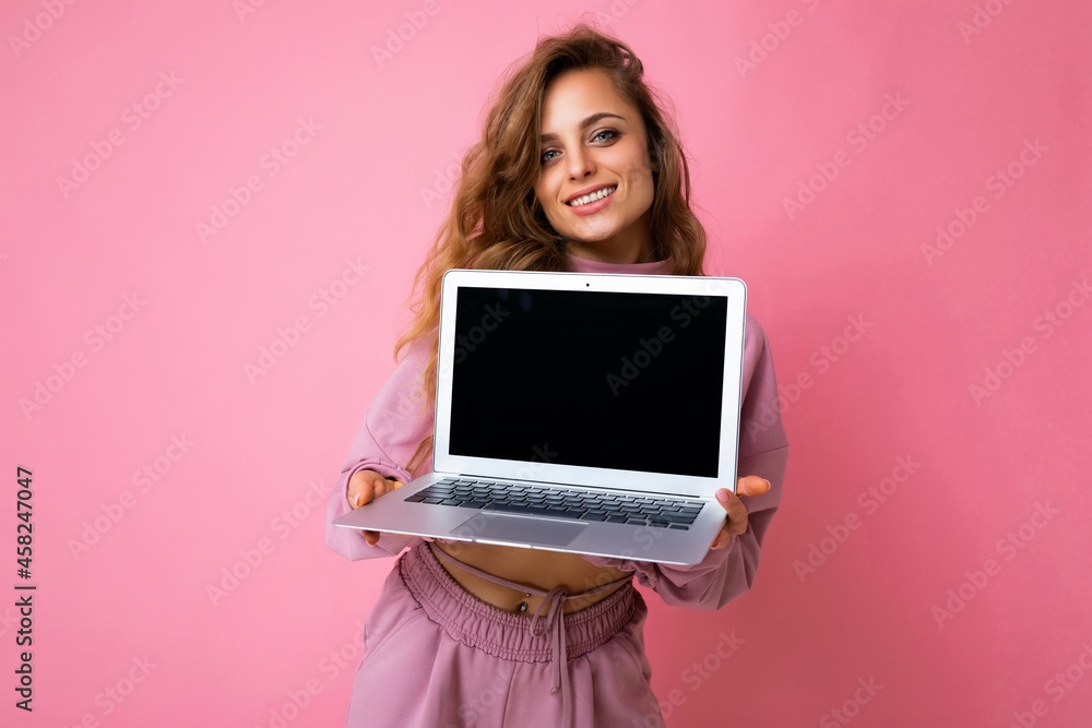 Naklejka premium Photo of beautiful charming happy smiling dark blond curly young woman lookind at camera holding computer laptop with empty monitor screen with mock up and copy space wearing pink hoodie isolated over