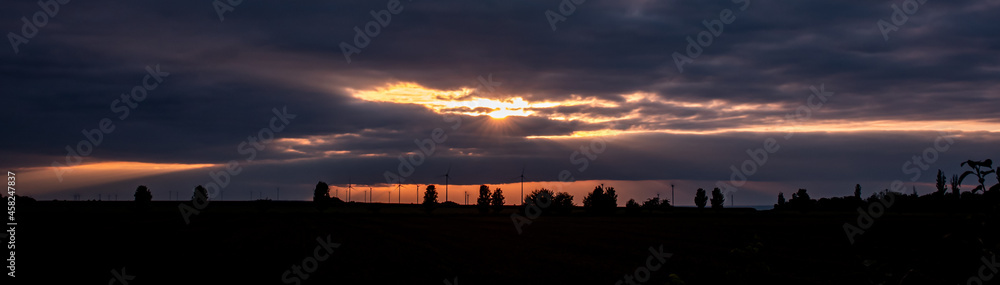 Silhouette of wind turbines and trees at the horizon during sunset. Dramatic orange glowing sky with a lot of clouds, sunbeams shines through the clouds.
