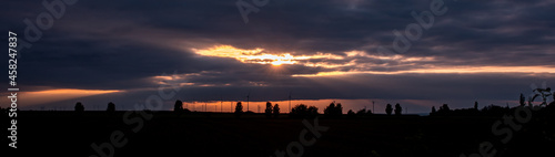 Silhouette of wind turbines and trees at the horizon during sunset. Dramatic orange glowing sky with a lot of clouds, sunbeams shines through the clouds. © Westlight