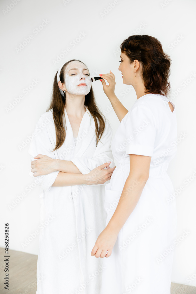 caucasian girl in a white coat with a white mask on her face. the cosmetologist applies the mask to the face with a brush. Facial peeling mask, spa beauty treatments, skin care. caucasian girl in a white coat with a white mask on her face. the cosmetologist applies the mask to the face with a brush. Facial peeling mask, spa beauty treatments, skin care.